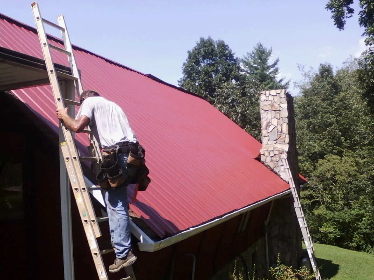 Roofer on a ladder beside a red metal roof and stone chimney — Soffit & Fascia Repair by Roofing Brothers in Salt Point